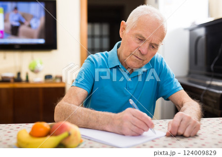 Serious older man sitting at table writing letter or filling some paper form 124009829