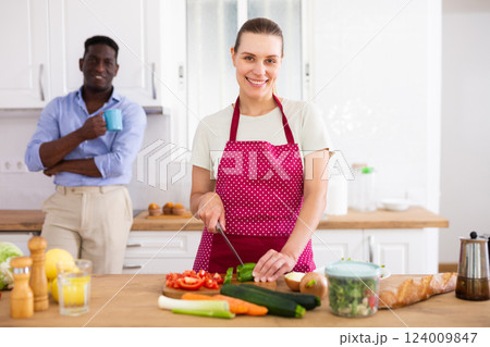Happy young woman cooking in home kitchen on background with smiling husband Happy young woman cooking in home kitchen on background with smiling husband 124009847
