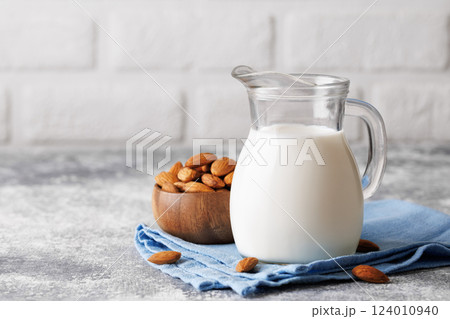 Glass milk jug resting on blue napkin beside wooden bowl filled with almonds, gray kitchen table featuring white brick wall backdrop, highlighting nutritious beverage 124010940