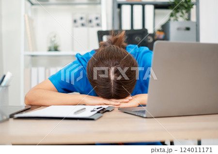 Tired healthcare professional in blue scrubs resting head on desk, surrounded by laptop and paperwork in a contemporary office setting. 124011171