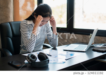 Young woman stressed over financial documents at office desk with laptop and headphones, in a modern workspace. 124011192