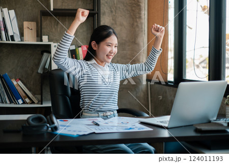 A young woman joyfully celebrates a work achievement in a modern office setting, surrounded by documents and a laptop. 124011193