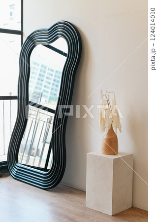 stylish mirror with wavy frame beside minimalist vase and dried leaves. modern decor enhances room aesthetic appeal 124014010