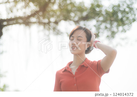 Asian woman in red shirt smiles with her eyes closed, enjoying sunlight in serene outdoor setting 124014372