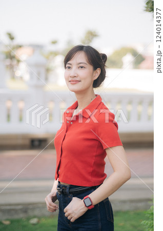 Asian young woman wearing red shirt and smartwatch stands outdoors, smiling confidently in park setting with blurred background 124014377