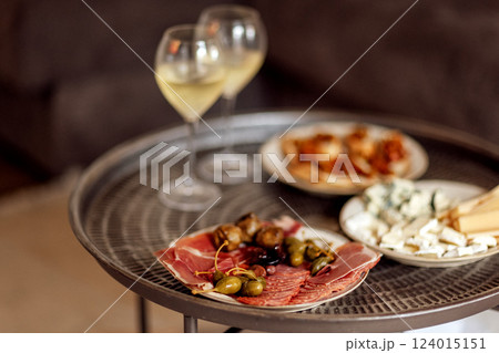 Two glasses of cool champagne and plates with various snacks on a round metal coffee table. White plates with slices of ham, salami, capers, olives and cheese. Two glasses of cool champagne and plates with various snacks on a round metal coffee table. White plates with slices of ham, salami, capers, olives and cheese. 124015151