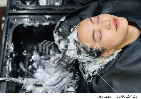 Asian Woman relaxing while getting her hair washed at salon, foam and water visible 124015423