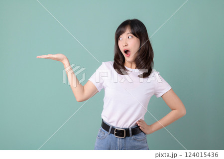 surprised young Asian woman in white t shirt and jeans gestures with her hand against green background 124015436