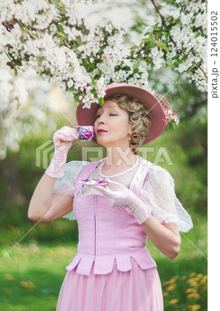 Happy natural adult mature woman of 60 years old in 18th century style pink hat, retro dress, gloves drinks tea in blossoming white apple orchard on summer day Happy natural adult mature woman of 60 years old in 18th century style pink hat, retro dress, gloves drinks tea in blossoming white apple orchard on summer day 124015502