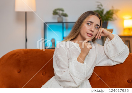 Thoughtful young woman in dress touching chin thinking about where to go for weekend sitting on sofa 124017926