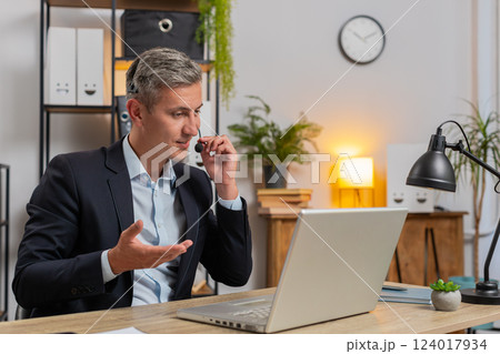 Confident young businessman in headset talking on video call using laptop computer at office desk Confident young businessman in headset talking on video call using laptop computer at office desk 124017934