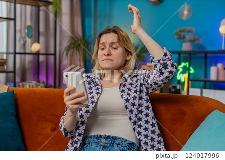 Shocked sad young woman using smartphone reading negative message feels annoyed sitting on sofa 124017996