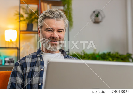 Smiling middle-aged mature man typing on laptop browsing internet using apps sitting at home sofa Smiling middle-aged mature man typing on laptop browsing internet using apps sitting at home sofa 124018025