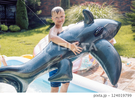 A little wet boy with an inflatable dolphin on the terrace of the house. A cute smiling child in the backyard pool. 124019799