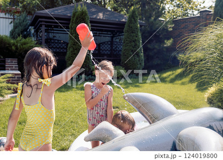 Small children splash water and have fun in the backyard of the house. Cute girls in swimsuits are doused with water and laughing. 124019800