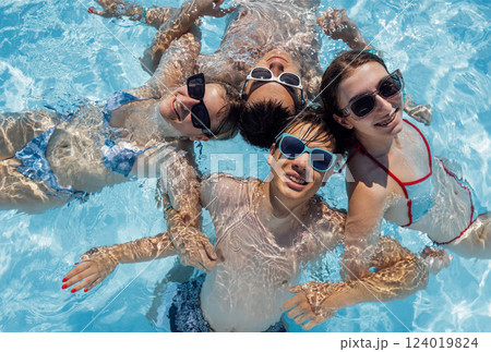 Smiling teenagers in swimsuits and sunglasses swim in the pool. A joyful group of friends is having fun in the clear water. Cute boys and girls are enjoying their holidays together. 124019824