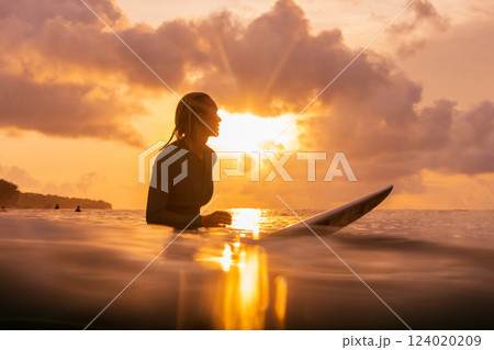 Attractive surf girl sitting on surfboard in ocean with sunset 124020209