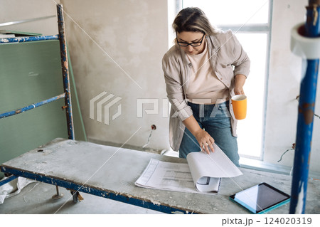 Young female designer checks renovation and house plan. Attractive woman in glasses and casual clothes drinks coffee and examines construction documentation. Young female designer checks renovation and house plan. Attractive woman in glasses and casual clothes drinks coffee and examines construction documentation. 124020319