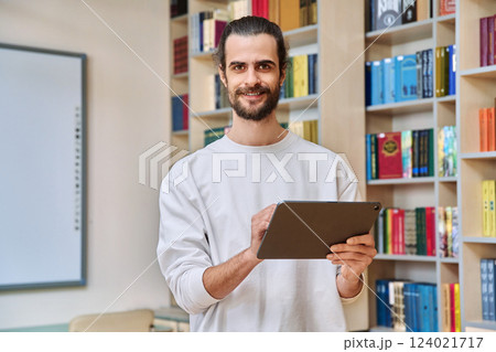 Young handsome man with digital tablet looking at camera inside library office 124021717