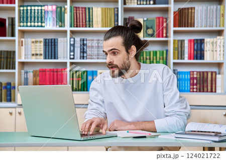 Young male teacher working in library sitting at desk with laptop books 124021722