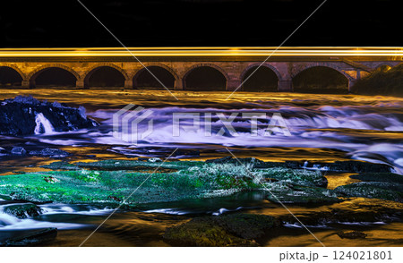 Flowing river under illuminated bridge at night with vibrant reflections and smooth water textures Flowing river under illuminated bridge at night with vibrant reflections and smooth water textures 124021801