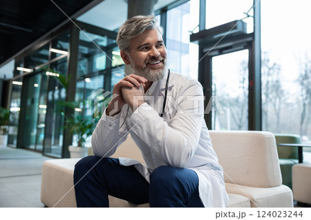Smiling doctor in lab coat sitting on the sofa in a clinic hall Smiling doctor in lab coat sitting on the sofa in a clinic hall 124023244