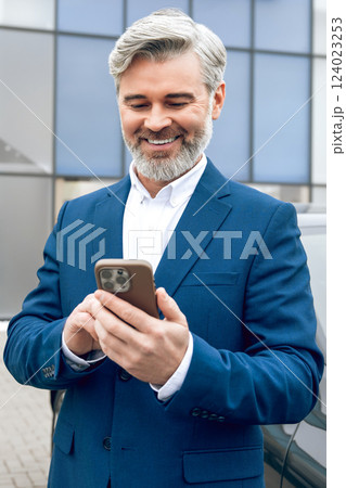 Car dealership worker in suit using smart phone and smiling while standing near car outdoor 124023253