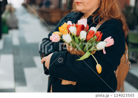 Beautiful young woman with spring tulips flowers bouquet walking at city street. Happy girl posing outdoors. Spring portrait of pretty female on urban background. High quality photo. 124023505