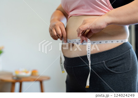 Overweight woman measuring her waist with a tape measure, focusing on weight loss progress at home Overweight woman measuring her waist with a tape measure, focusing on weight loss progress at home 124024249