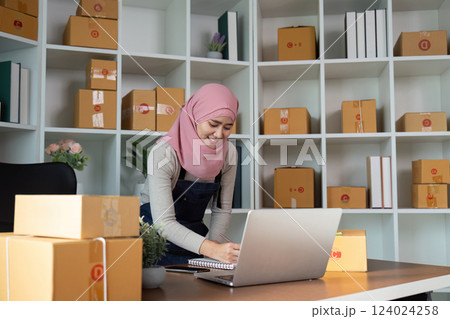 Muslim woman organizing online sales while writing notes at her desk, showcasing her e-commerce business in a well-arranged workspace. 124024258