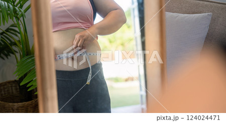 Woman measuring her waist with a tape measure while examining her reflection, focusing on body image goals 124024471