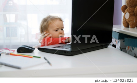 Cute baby girl sits at her desk typing on a laptop, surrounded by coloring pens and a notebook. Young freelancer working in a cozy space 124028823