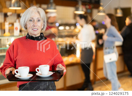 Senior woman visitor in self-service cafeteria carries tray with two cups of coffee. 124029175
