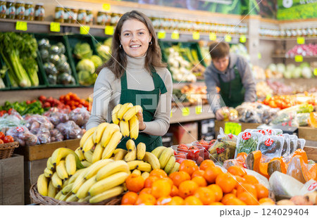 Positive female seller in apron displaying assortment of bananas at supermarket Positive female seller in apron displaying assortment of bananas at supermarket 124029404