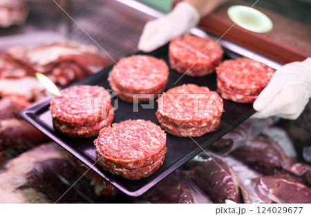 Tray with burger patties held in hands by seller against background of showcase of butcher shop 124029677