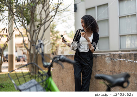 Outdoor Connectivity and Communication. A smiling young woman enjoys her coffee while using her phone, with a bicycle close by in a park setting. 124036628