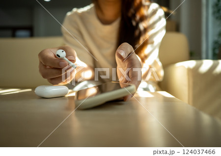 Young woman holding a wireless earbud and reaching for her smartphone Young woman holding a wireless earbud and reaching for her smartphone 124037466