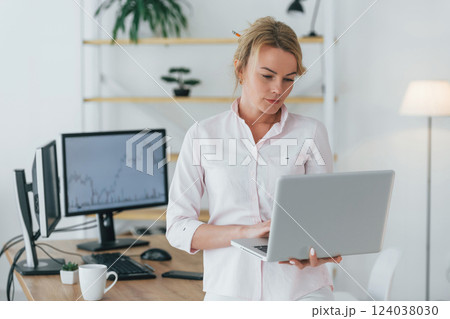 Standing with laptop in hands. Female stock broker in formal wear is working in the office Standing with laptop in hands. Female stock broker in formal wear is working in the office 124038030
