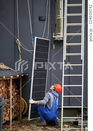 Men installers installing solar panel system on roof of house. Workers in helmets lifting up photovoltaic solar module with help of ropes outdoors. Concept of alternative and renewable energy. Men installers installing solar panel system on roof of house. Workers in helmets lifting up photovoltaic solar module with help of ropes outdoors. Concept of alternative and renewable energy. 124039742