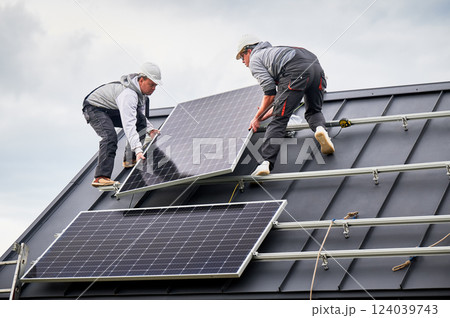 Installers installing solar panel system on roof of house. Men workers in helmets carrying photovoltaic solar module outdoors. Concept of alternative and renewable energy. 124039743