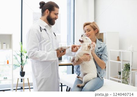 Man veterinarian with young adult woman owner examining white dog at modern clinic. Smiling doctor providing care. Concept of pet health, animal care, veterinary service, friendship. 124040714