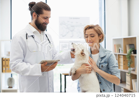 Man veterinarian with young adult woman owner examining white dog at modern clinic. Smiling doctor providing care. Concept of pet health, animal care, veterinary service, friendship. 124040715