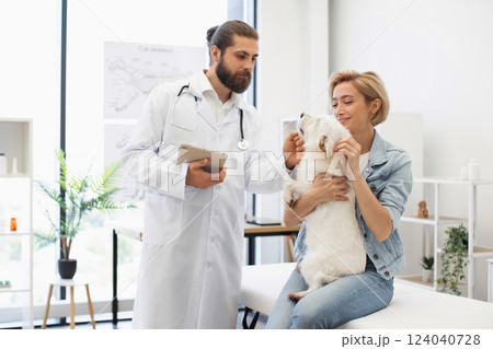 Veterinarian conducting routine checkup shows professional care. Female owner holding dog loving manner. Modern veterinary clinic setting. 124040728