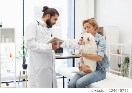 Male veterinarian in white coat examines dog held by young adult woman at clinic. Modern light interior. Interaction communicates pet healthcare, caring relationship, professional service. Male veterinarian in white coat examines dog held by young adult woman at clinic. Modern light interior. Interaction communicates pet healthcare, caring relationship, professional service. 124040732