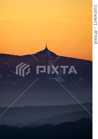 As the sun sets, a stunning silhouette emerges on Jested ridge, casting a serene outline against the orange and purple sky. The surrounding landscape fades into misty hills, creating a tranquil vista. 124041651