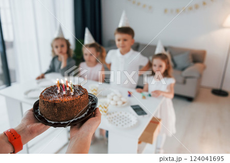 Woman holding cake with candles. Celebrating birthday. Group of children is together at home at daytime Woman holding cake with candles. Celebrating birthday. Group of children is together at home at daytime 124041695
