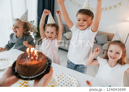Woman holding cake with candles. Celebrating birthday. Group of children is together at home at daytime Woman holding cake with candles. Celebrating birthday. Group of children is together at home at daytime 124041699