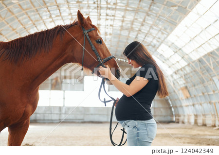 Side view. Beautiful pregnant woman in casual clothes is with the horse on a stable 124042349