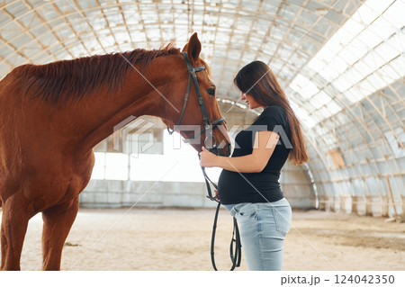 Side view. Beautiful pregnant woman in casual clothes is with the horse on a stable 124042350