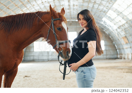 Looking at the camera. Beautiful pregnant woman in casual clothes is with the horse on a stable 124042352